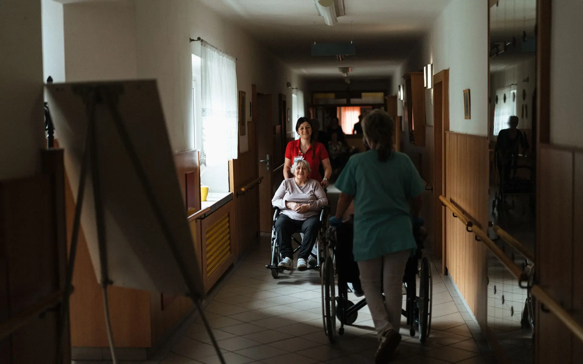 Caregiver walking with a senior woman in a wheelchair through a care community hallway